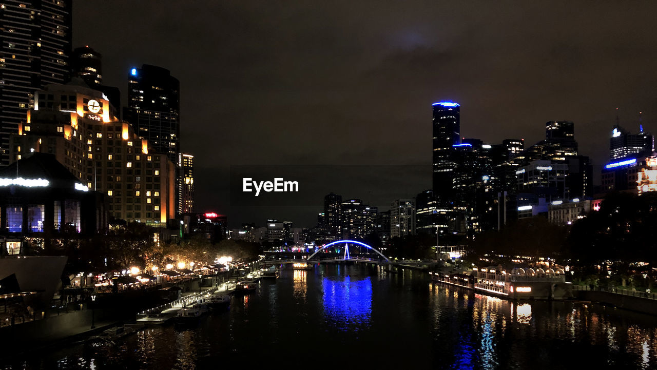 ILLUMINATED BRIDGE OVER RIVER BY BUILDINGS AGAINST SKY