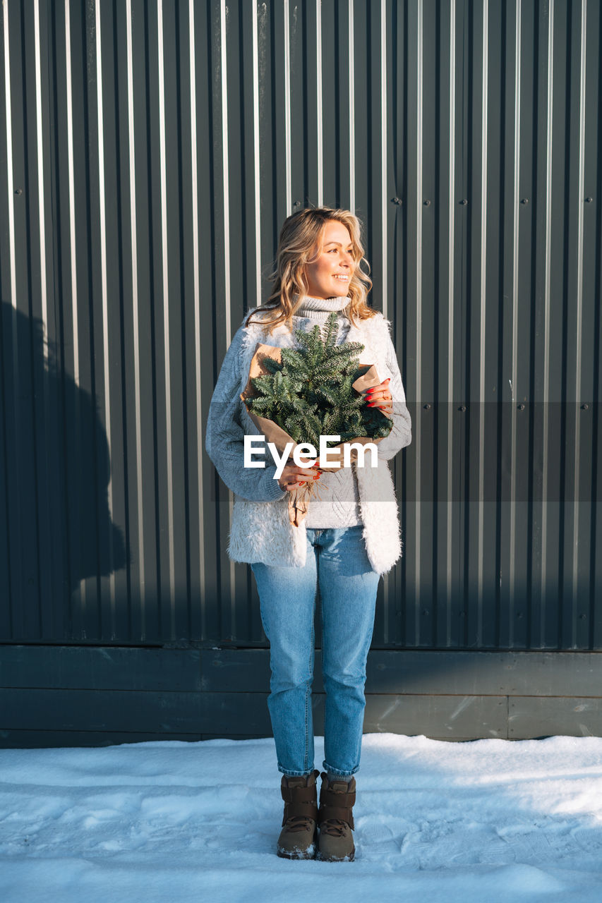 Portrait of woman in winter clothes with fir branches in hands against background of grey wall