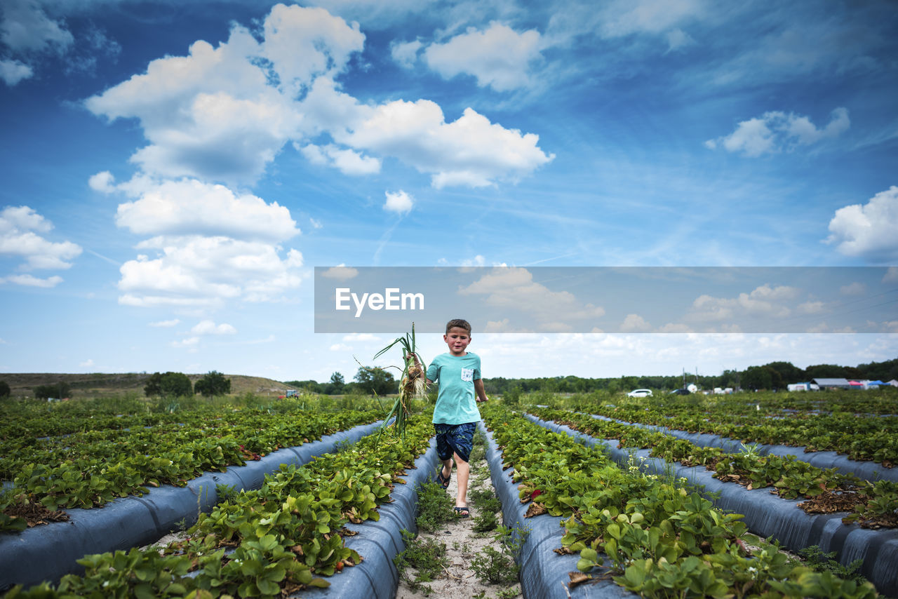 Boy holding root vegetables while running at organic farm against cloudy sky