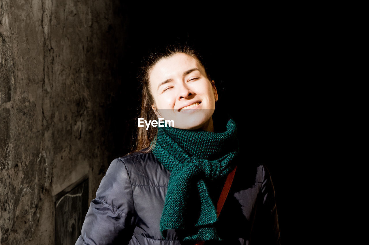 Young smiling woman wearing warm clothing by wall at night