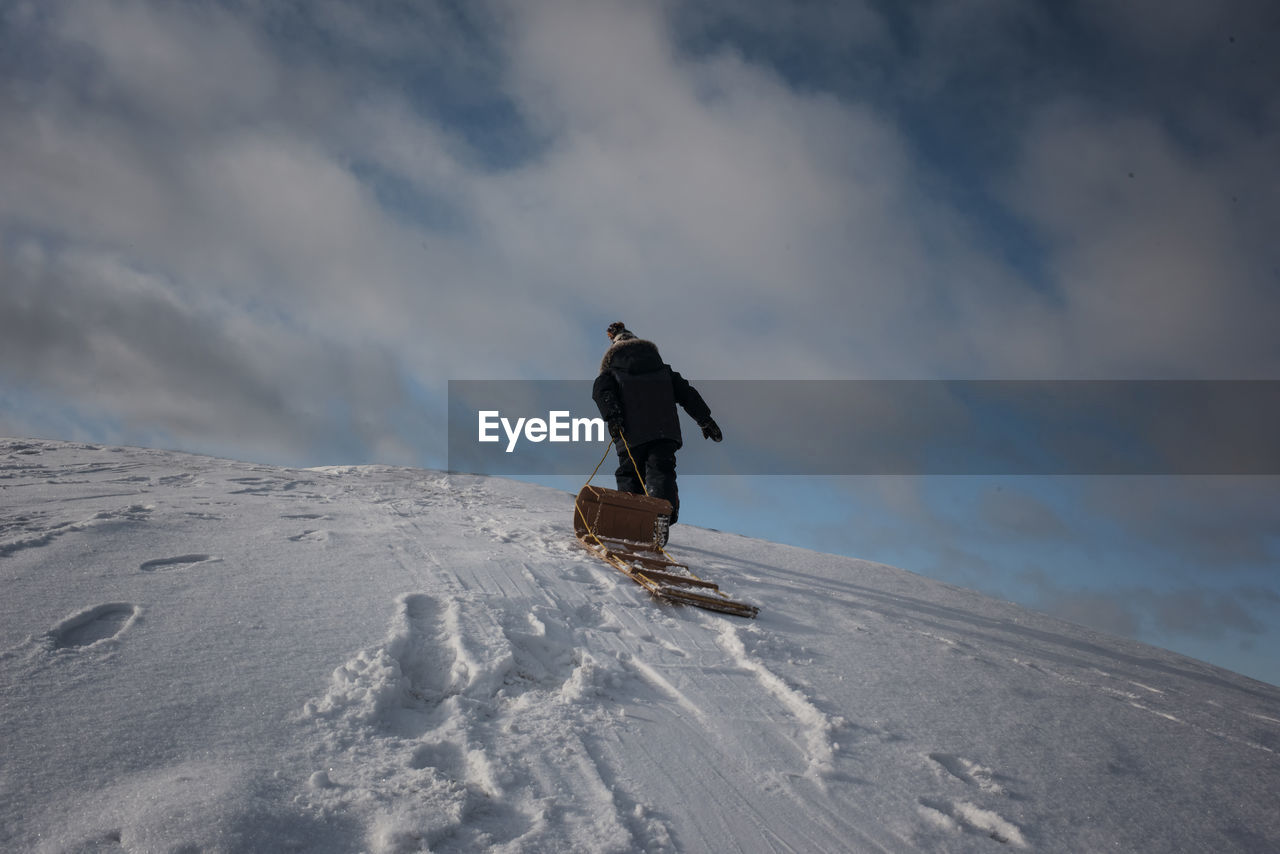 Low angle view of man pulling sled while walking on snow covered mountain against cloudy sky