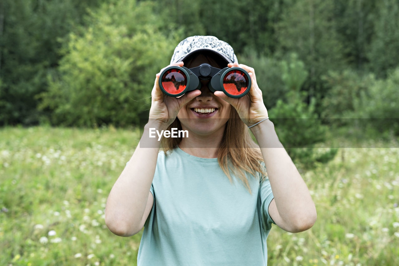 Happy young blonde woman bird watcher in cap looking through binoculars at cloudy sky in forest 