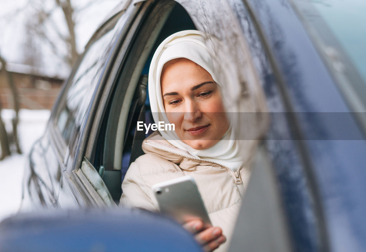 Beautiful smiling young muslim woman in headscarf in clothing using mobile in right-hand-drive car