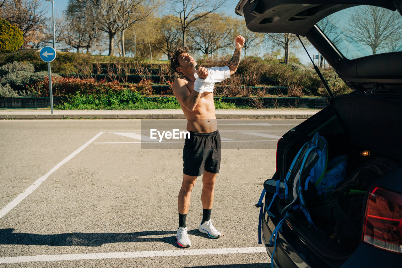 Masculine male athlete with tattoo putting on t shirt against automobile in parking on sunny day