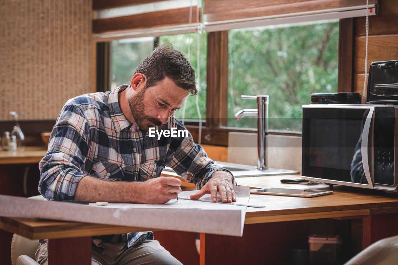 Serious male architect sitting at table in kitchen and drawing blueprint of building with pencil and ruler while working at home