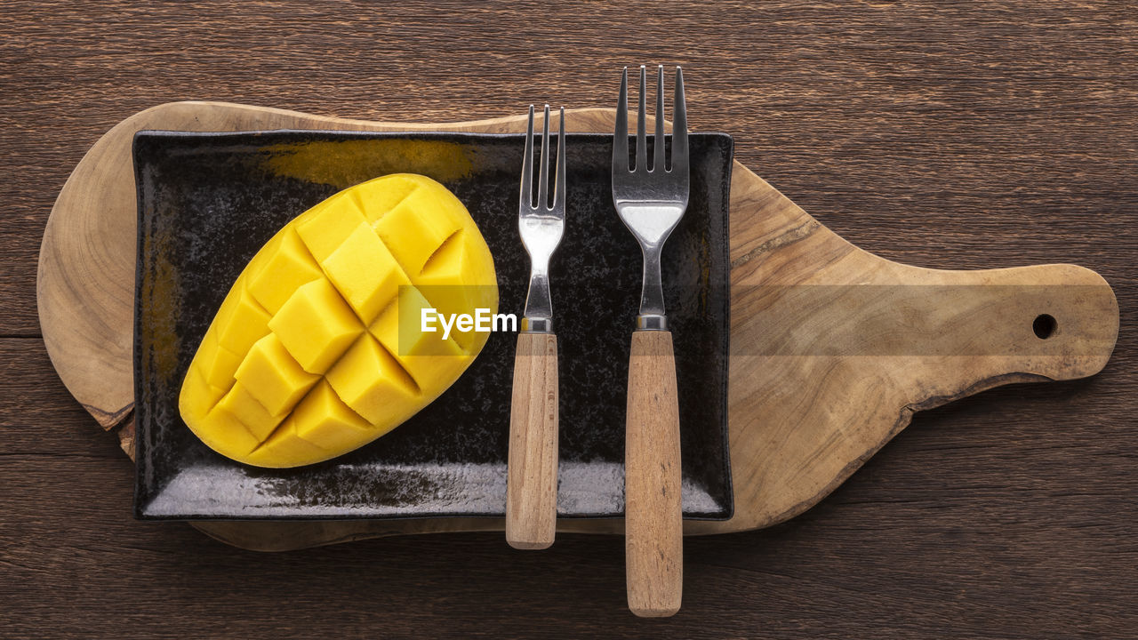 Tasty fresh yellow ripe mango with fork in black ceramic plate on olive wood cutting board, top view