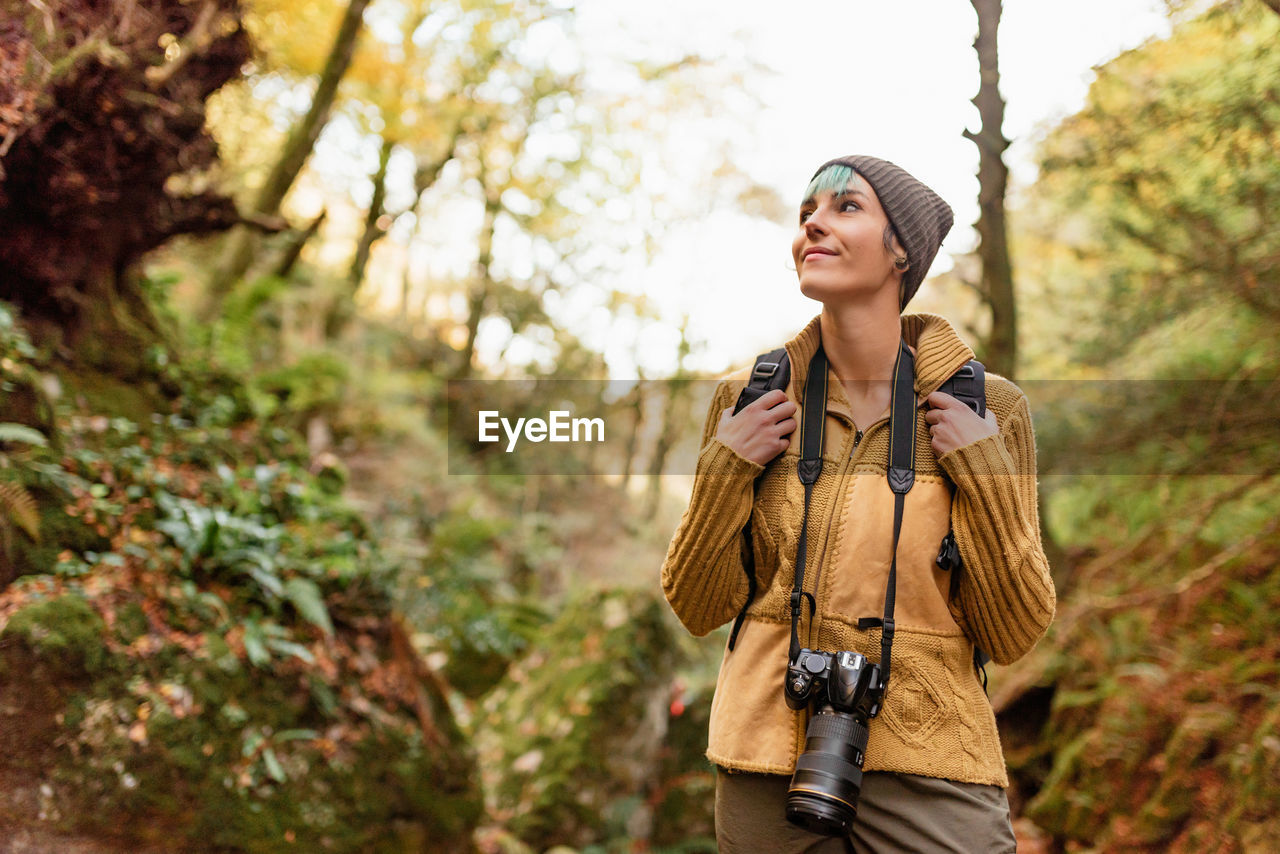 Low angle of traveling photographer standing in woods with photo camera and looking up while admiring nature during vacation