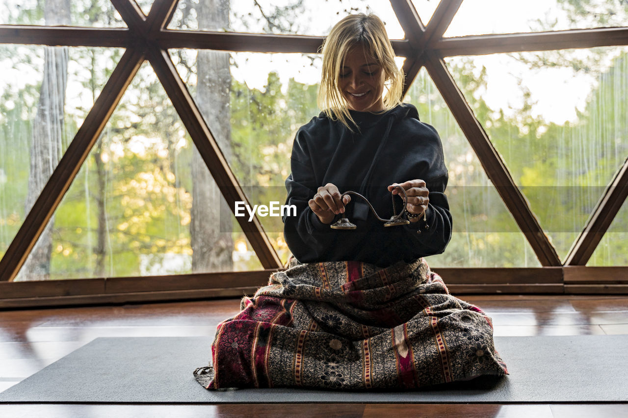 Smiling blond female yoga instructor holding equipment while sitting on mat against window in exercise class