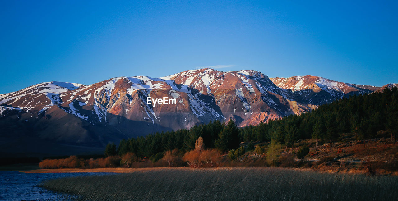 Calm countryside lake in front of mountains against clear blue sky