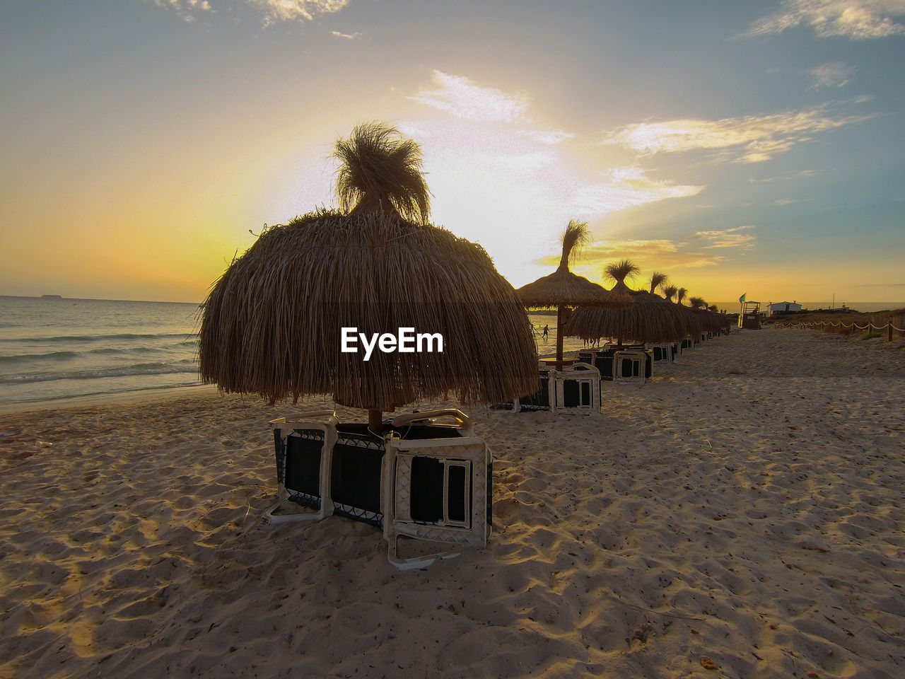 Beach umbrellas on sand with folded sunbeds, with blue sky and the sun setting. in mallorca, spain