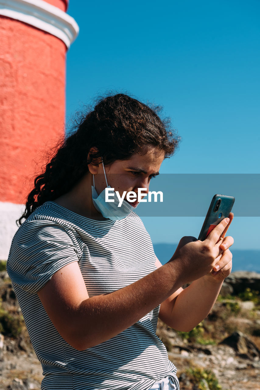 Young woman wearing face mask using mobile phone against sky