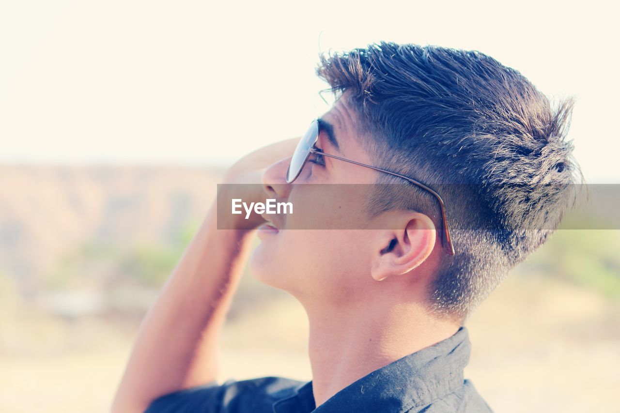 Side view of young man in sunglasses standing against sky