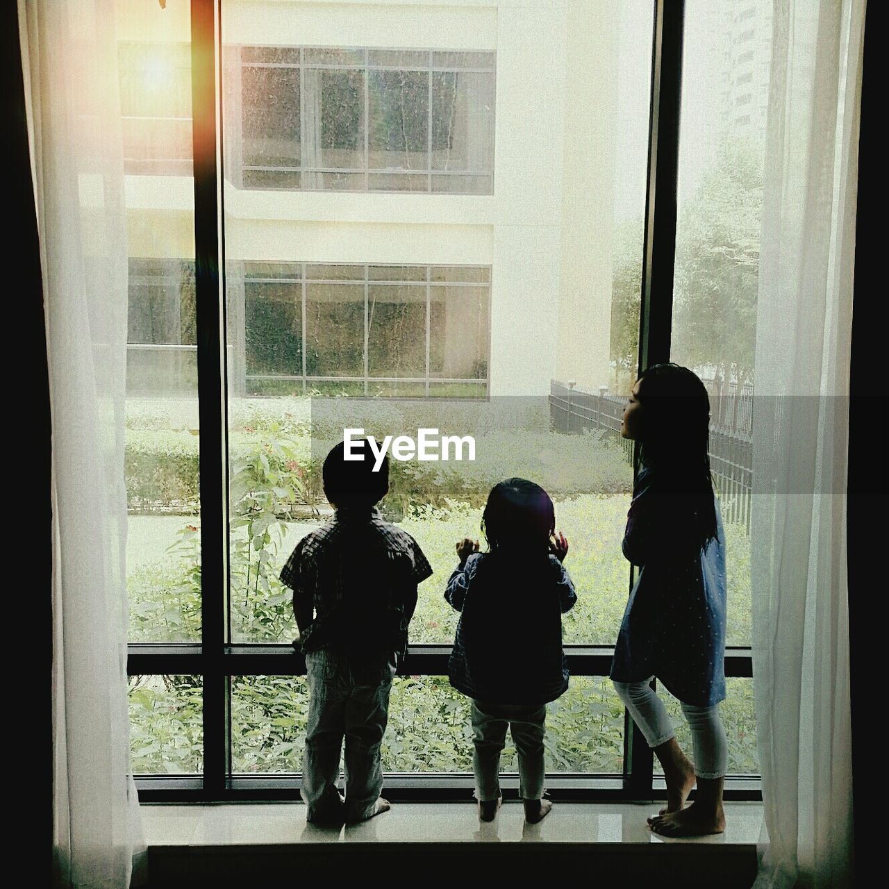 Siblings looking through window while standing at home