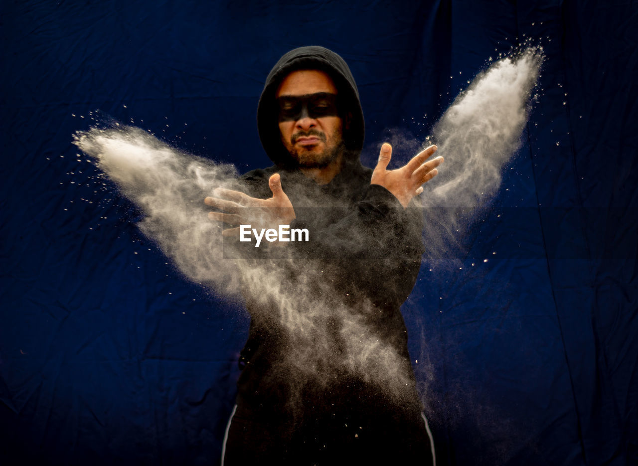 Man wearing hood with face paint throwing powder against backdrop