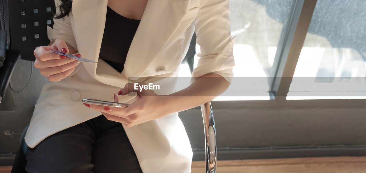 MIDSECTION OF WOMAN HOLDING SMART PHONE WHILE STANDING ON WINDOW