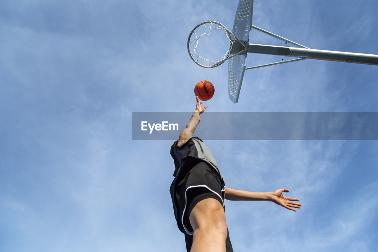 From below of anonymous sportive male throwing ball into basketball hoop while playing game against blue sky on sunny summer day