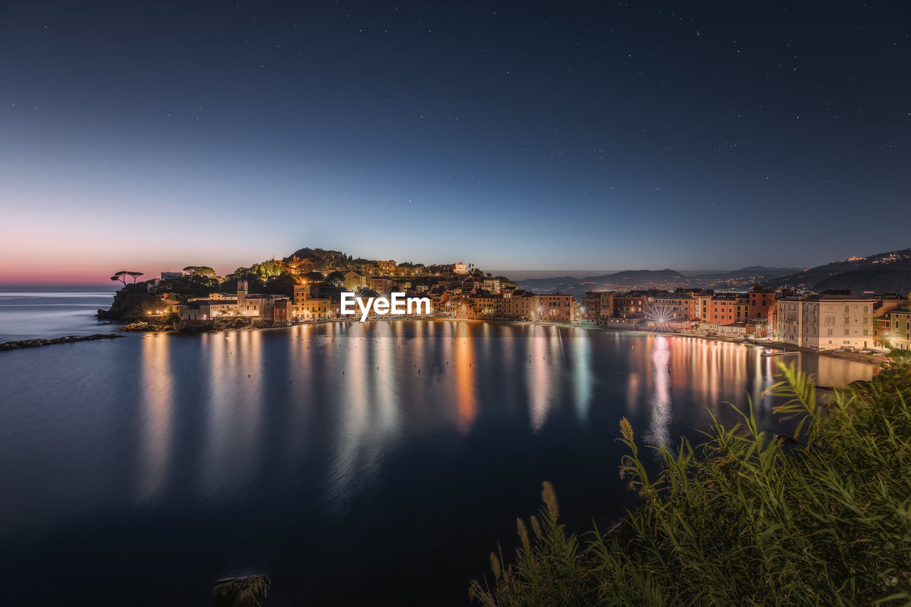 Illuminated buildings by lake against sky in city at night, bay of silence, sestri levante, italy 