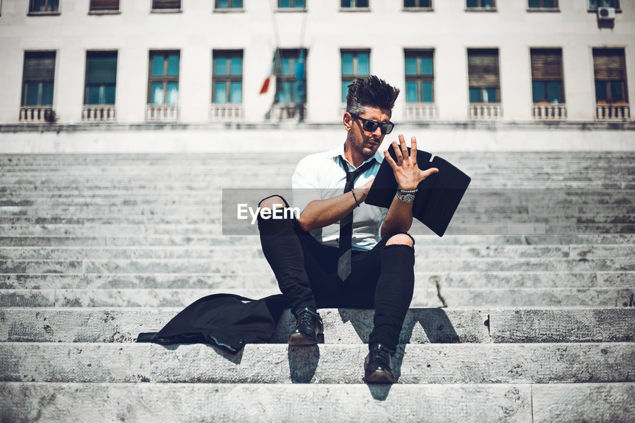 Full length of man using digital tablet while sitting on staircase in city