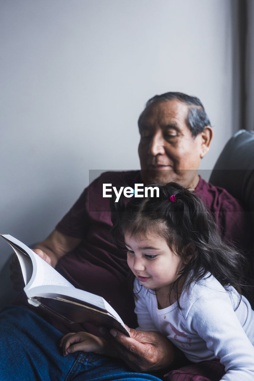 Senior ethnic grandfather reading interesting story in book for cute little girl while relaxing together on sofa at home