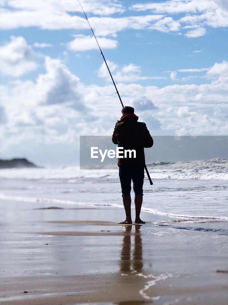 Rear view of man with fishing rod standing at beach against cloudy sky