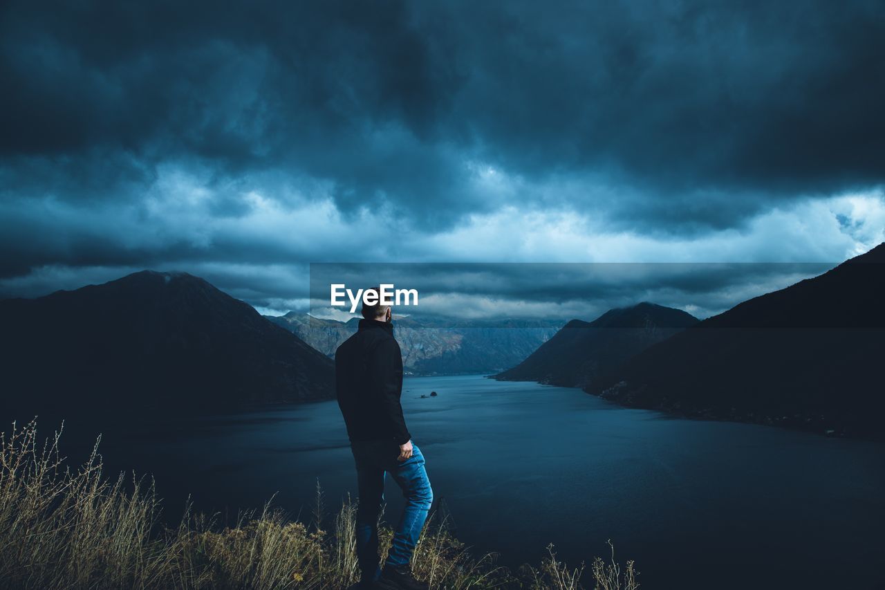 Rear view of man standing on mountain by river against cloudy sky