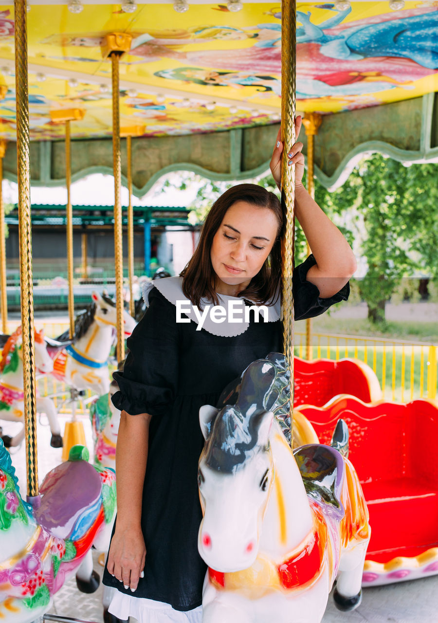 Beautiful brunette girl in a black dress poses on a bright carousel with horses