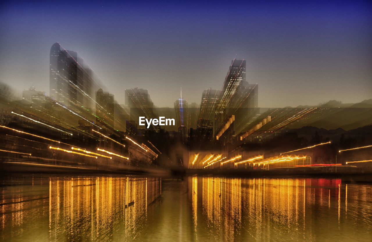 ILLUMINATED CITY BUILDINGS BY RIVER AGAINST SKY