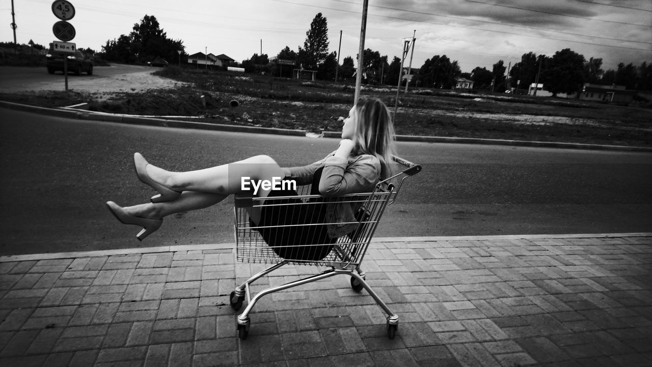 Side view of woman sitting in shopping cart on footpath