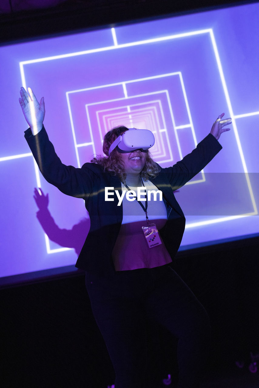 Happy businesswoman wearing vr glasses standing with arms raised in convention center