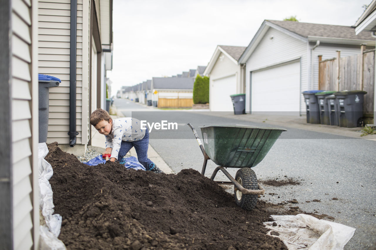 Young boy scooping pile of soil into wheelbarrow in back alley