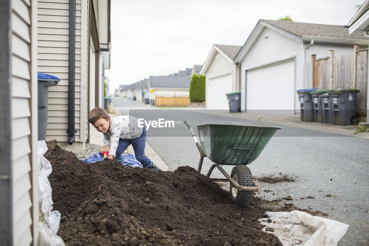 Young boy scooping pile of soil into wheelbarrow in back alley