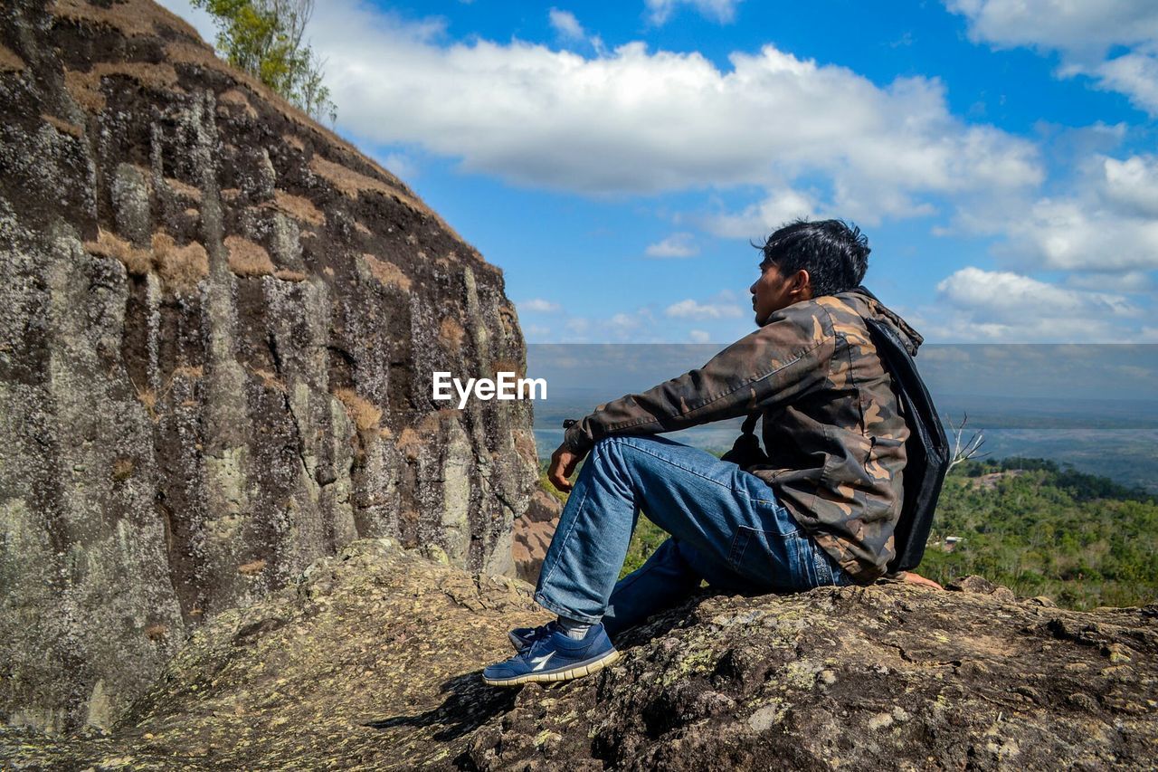 REAR VIEW OF MAN SITTING ON ROCK AGAINST SKY