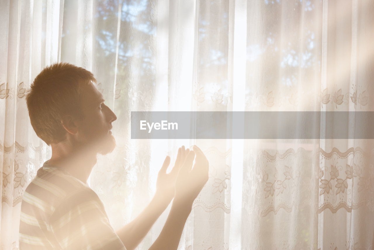 SIDE VIEW OF YOUNG WOMAN LOOKING THROUGH WINDOW AT CURTAIN