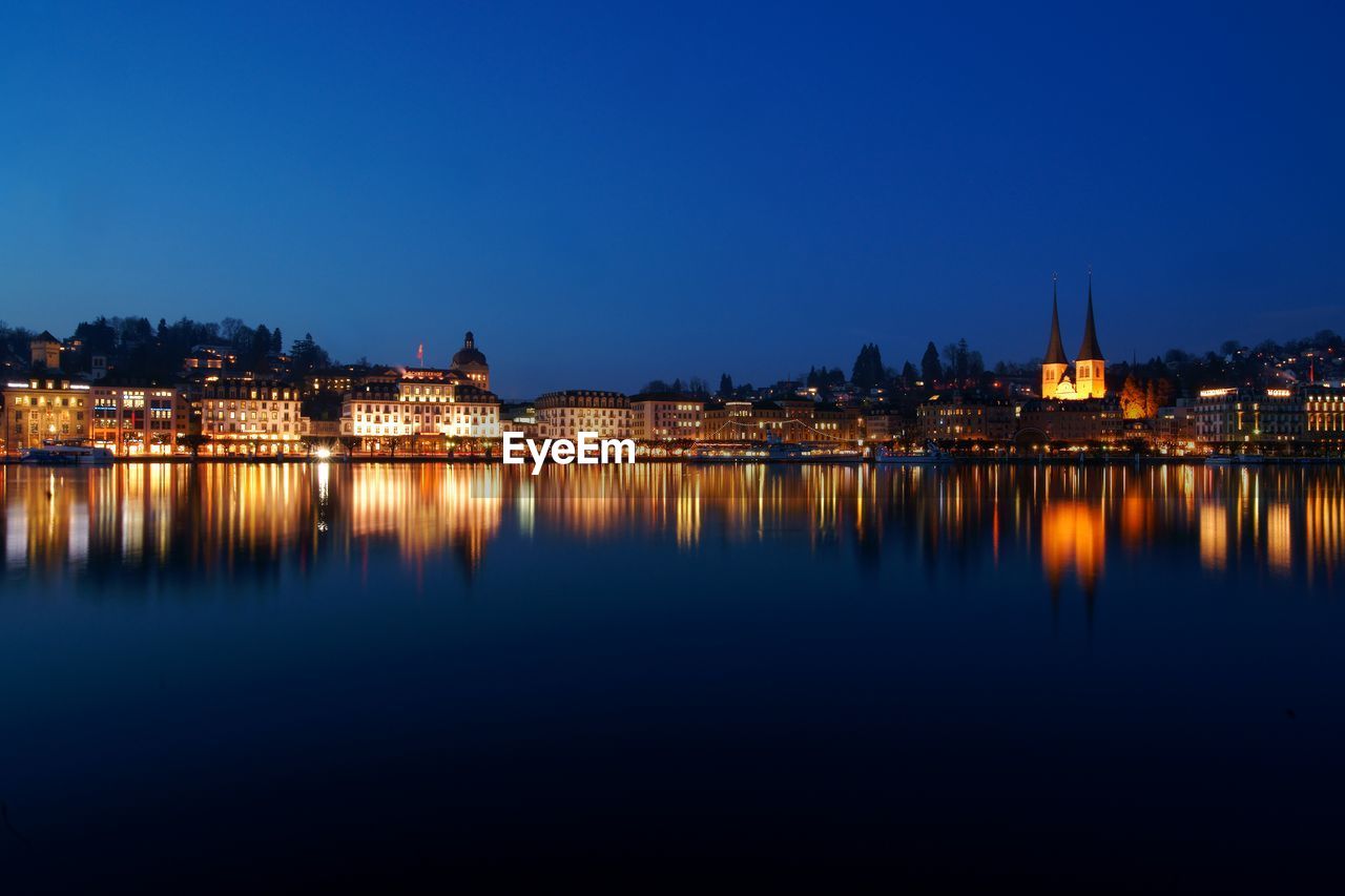 Illuminated buildings by river against sky at dusk