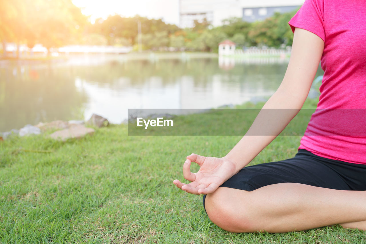Midsection of woman practicing yoga on grass field by lake in park