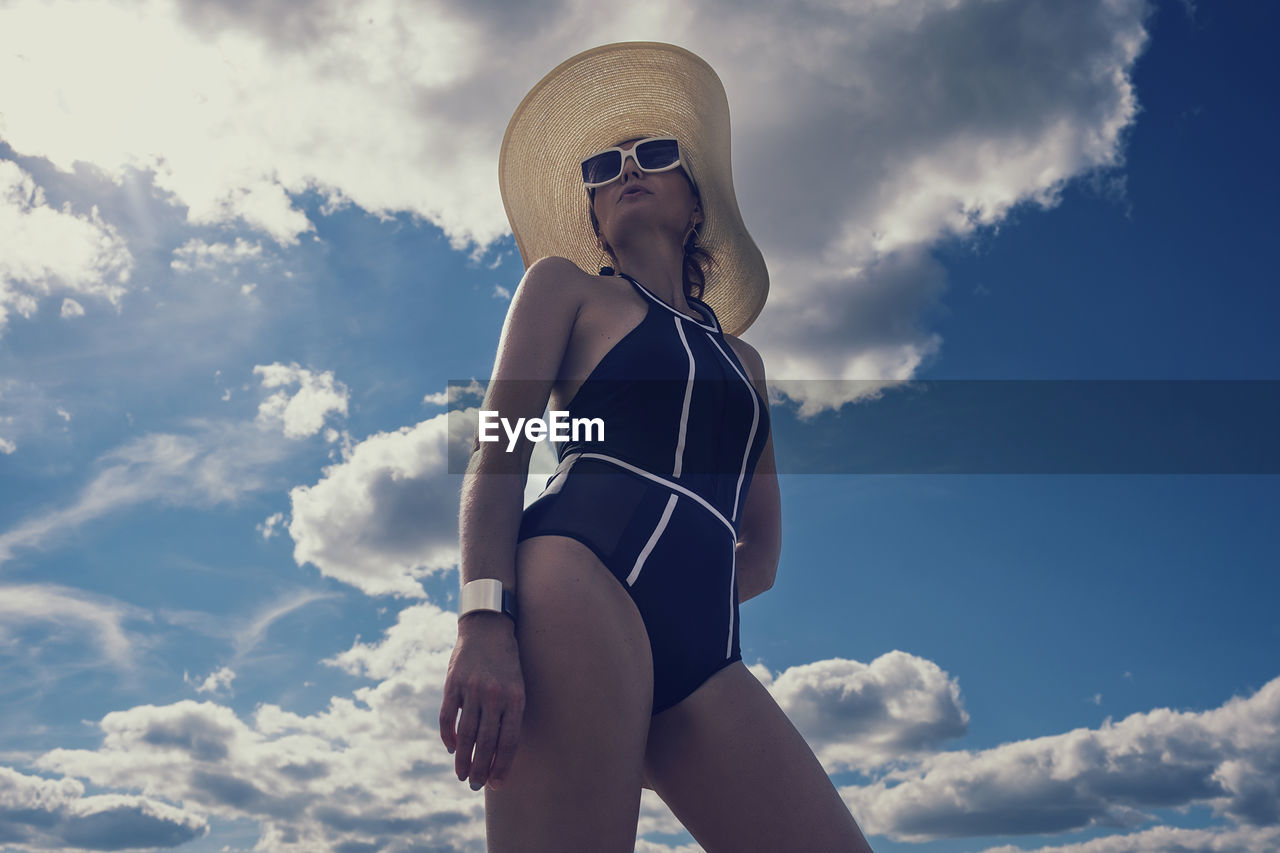 A portrait of a woman in a swimsuit, hat and sunglasses in summer on the riverbank against the sky