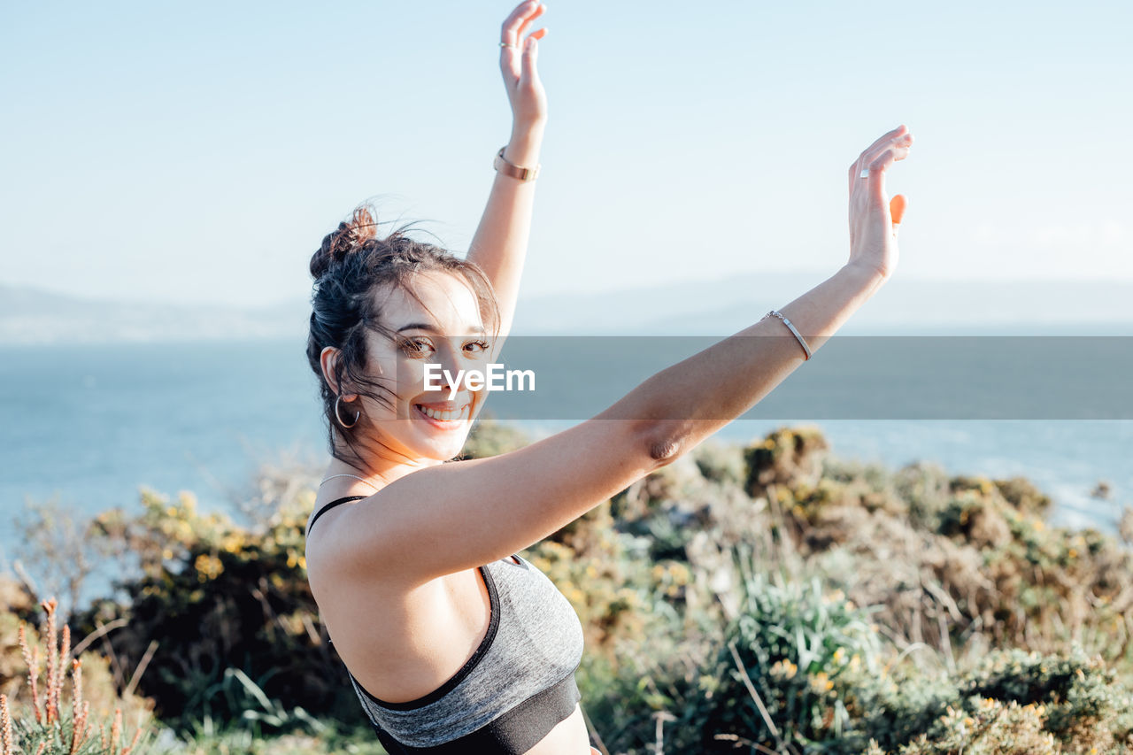 Portrait of young woman with arms raised against sea