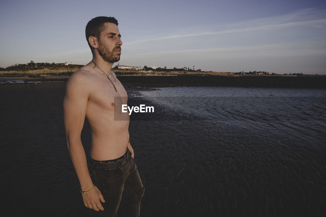Young shirtless man looking at sea against sky