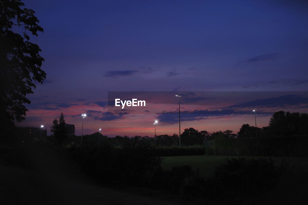 Scenic view of trees and floodlights over soccer pitches, against sky at dusk