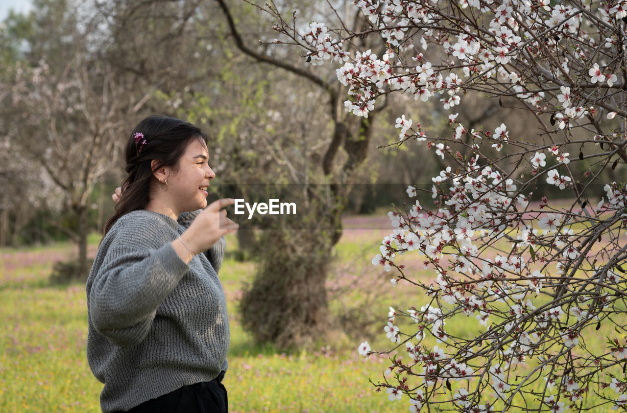 Side view of young woman standing by cherry blossom plants