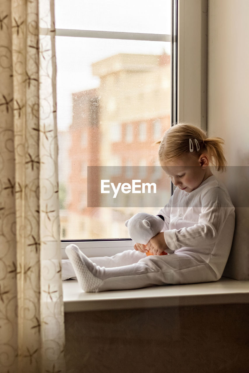 Full length of girl with stuffed toy sitting by window