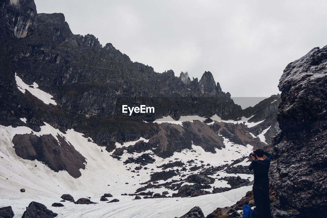 Rear view of man photographing amidst snow covered mountains against sky