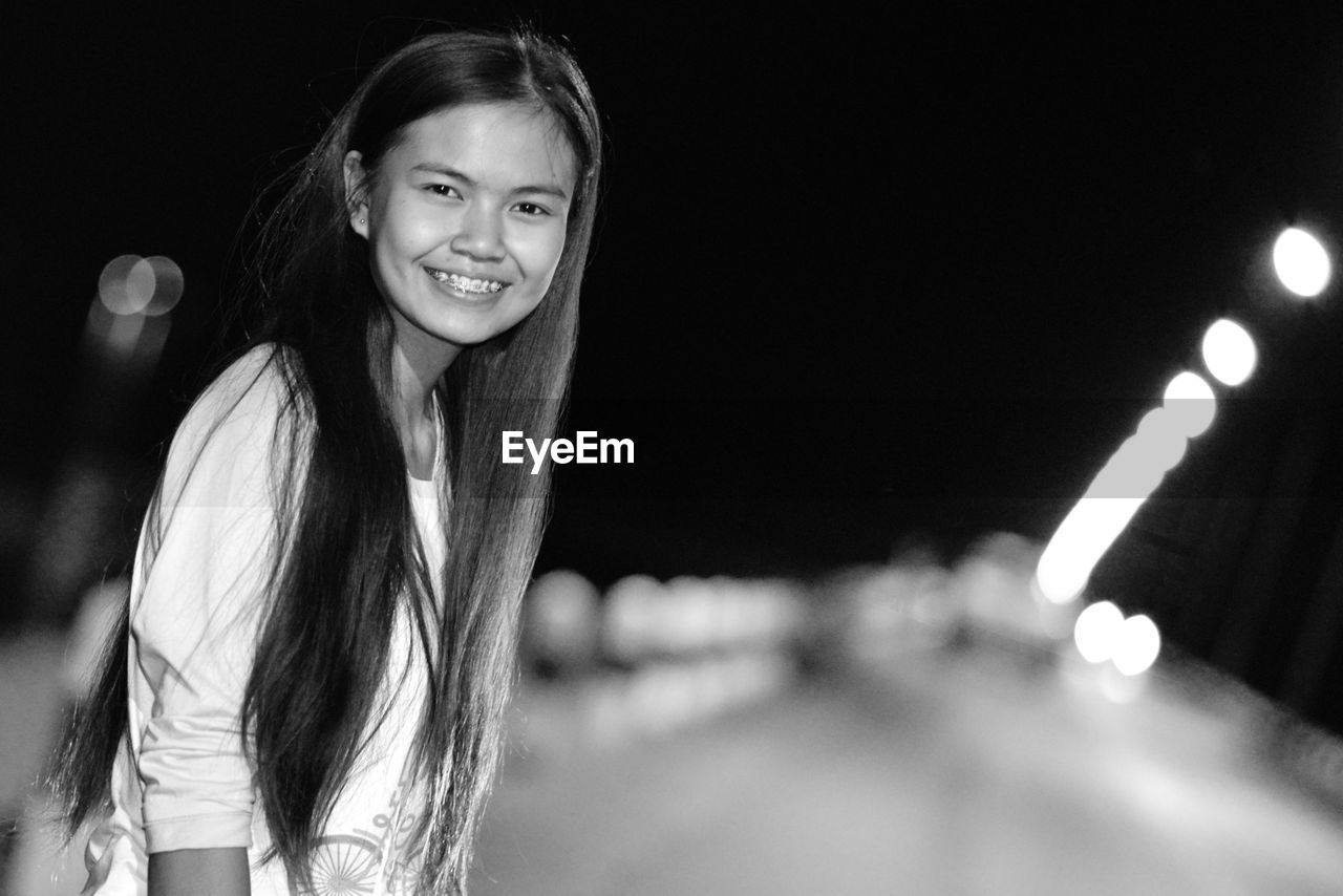 Portrait of happy young woman standing on footpath at night