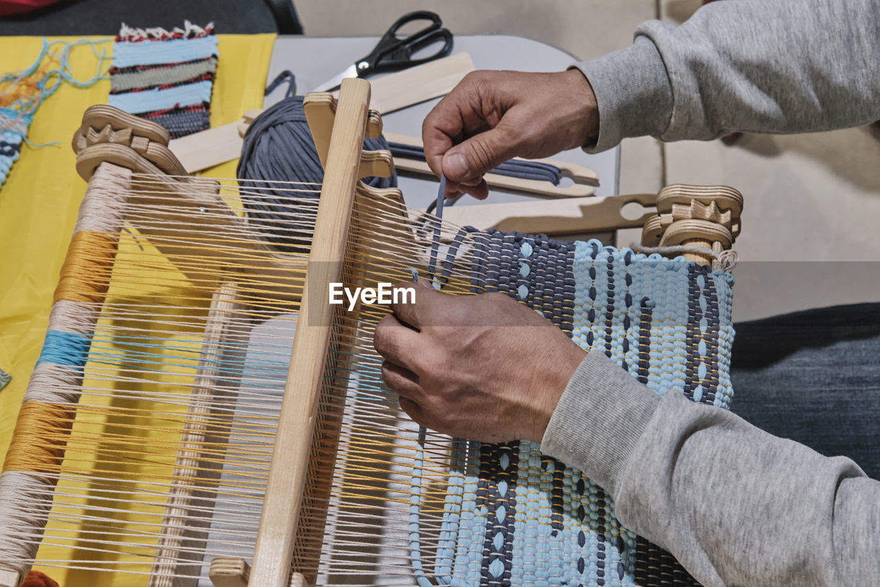 Hands of senior man weaving small rug with pattern on manual table loom.