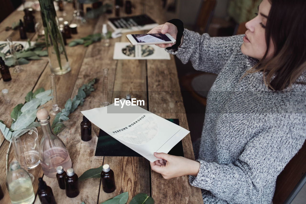 High angle view of mid adult female owner photographing brochure while sitting at table in workshop