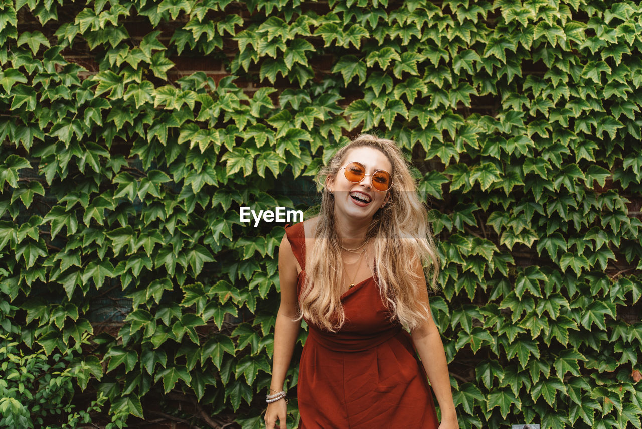 Cheerful young woman wearing sunglasses while standing against plants