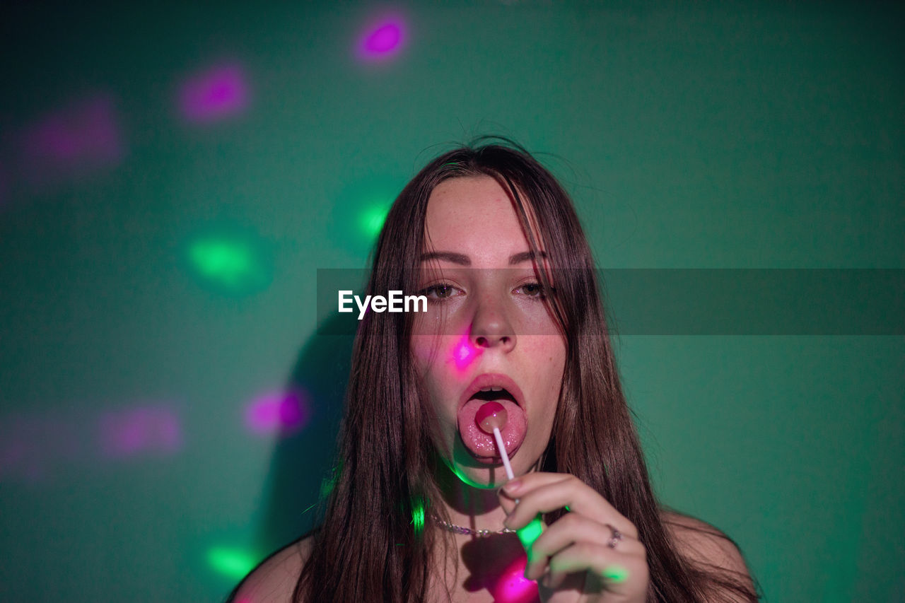 Portrait of young woman licking lollipop while lights falling on her against green background