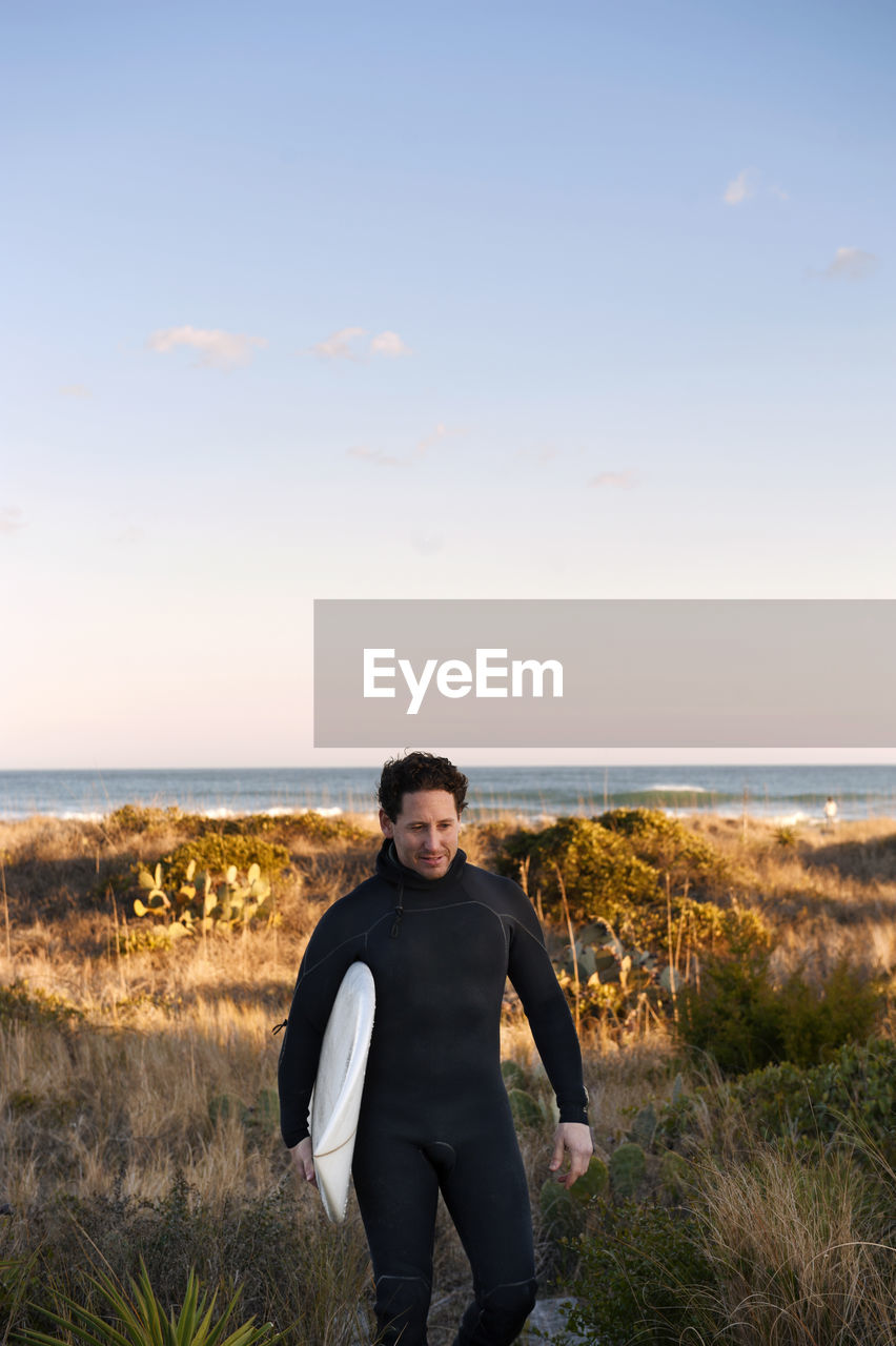 Surfer carrying surfboard while walking on grassy field against sky