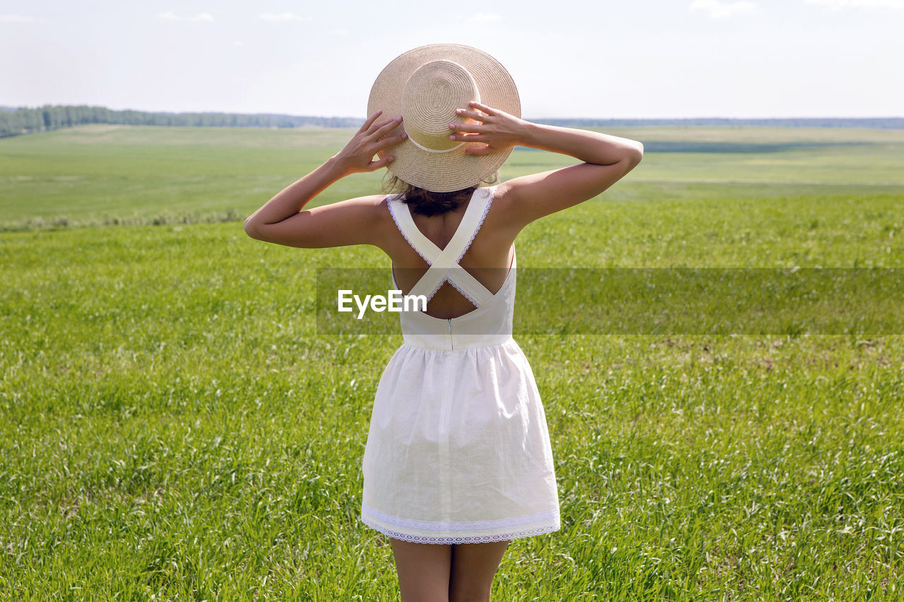 Young woman with long dark hair standing rear on green field. white dress and straw hat. 