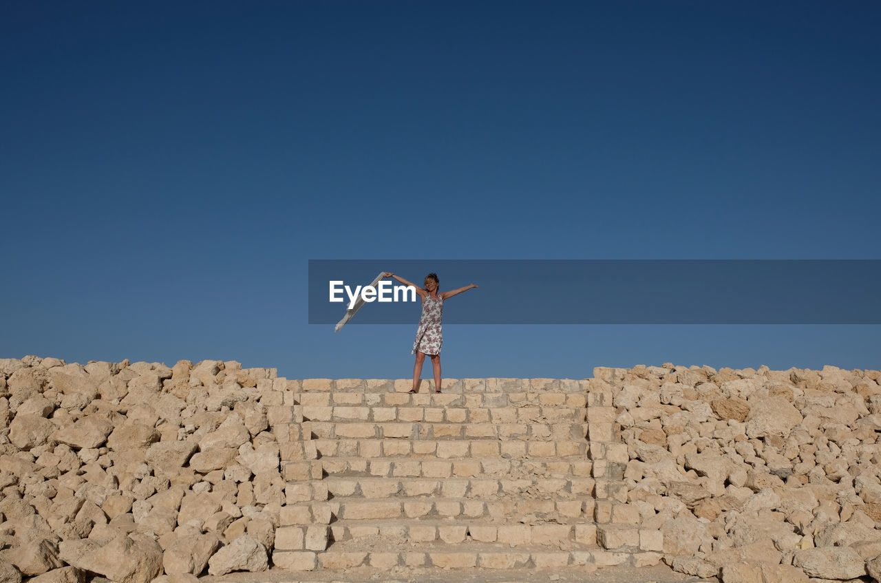 Woman with arms outstretched standing on steps against clear blue sky